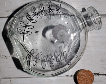 May include: A clear glass bottle with a black and white design of leaves and a crescent moon. The bottle has a cork stopper.