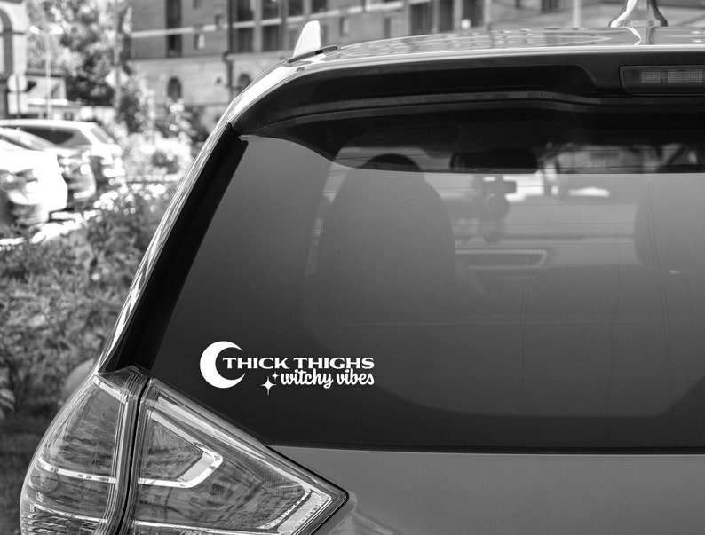 May include: A black car with a white vinyl sticker on the back window. The sticker says "Thick Thighs + Witchy Vibes" with a crescent moon and a star.