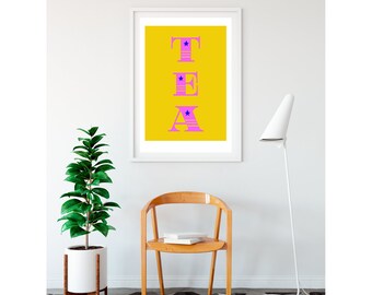 Op de afbeelding: Een witte ingelijste print met een gele achtergrond en het woord "TEA" in roze letters met blauwe sterren. De print hangt aan een witte muur boven een houten stoel en een witte staande lamp.