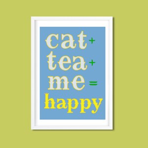Op de afbeelding: Een ingelijste kunstdruk met de tekst "cat + tea + me = happy" in een speels lettertype. De tekst is wit en geel op een lichtblauwe achtergrond, met groene plus- en isgelijktekens. De lijst is wit.