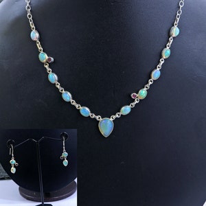 May include: A silver necklace with a teardrop-shaped opal pendant and smaller opal stones along the chain. The necklace also features small red gemstones. Matching earrings with opal and red gemstones are included.