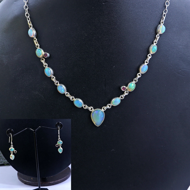 May include: A silver necklace with a teardrop-shaped opal pendant and smaller opal stones along the chain. The necklace also features small red gemstones. Matching earrings with opal and red gemstones are included.