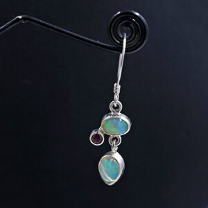 May include: Silver dangle earrings with three opal stones and one small red gemstone. The opal stones are oval and teardrop shaped.