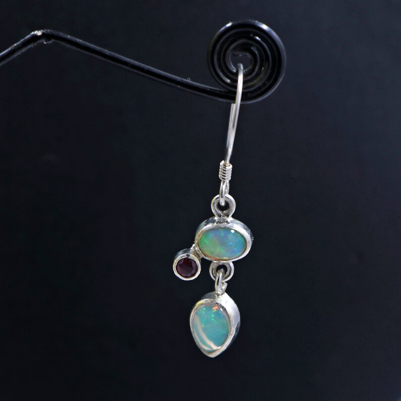 May include: Silver dangle earrings with three opal stones and one small red gemstone. The opal stones are oval and teardrop shaped.
