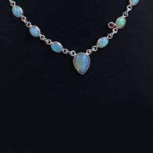May include: A silver necklace with a teardrop-shaped opal pendant and smaller opal stones along the chain. The necklace has a small red stone accent.