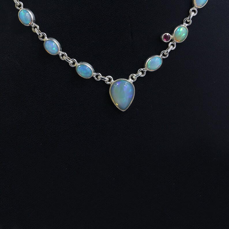 May include: A silver necklace with a teardrop-shaped opal pendant and smaller opal stones along the chain. The necklace has a small red stone accent.