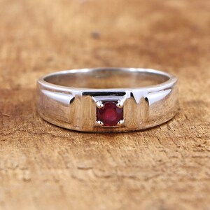 Può includere: Un anello a fascia d'argento con una singola pietra preziosa rossa incastonata al centro.