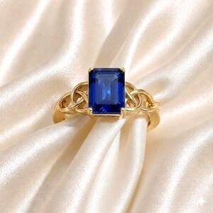 Puede incluir: Un anillo de oro con una gran piedra preciosa rectangular de color azul oscuro. El anillo tiene un diseño de nudo celta en los laterales. El anillo está sobre un fondo de tela sedosa de color crema.