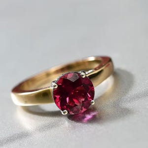 Peut inclure: Une bague en or avec une pierre précieuse ronde à facettes d'un rouge profond, fixée par un sertissage à quatre griffes. L'anneau est lisse et poli, présentant un design classique avec une pierre cramoisie vive comme point focal central.