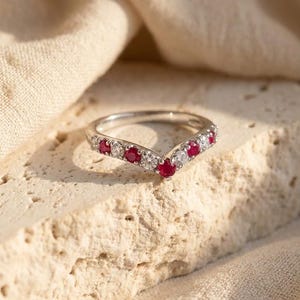 May include: A V-shaped silver-toned ring featuring an alternating pattern of round-cut red rubies and clear white stones set along the band, with a central ruby at the point of the V-shape, showcasing a polished metal finish and prong settings.