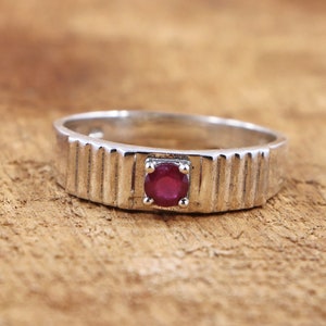 Puede incluir: Un anillo de plata con una piedra preciosa de rubí rojo engastada en el centro. La banda del anillo tiene un diseño texturizado y estriado.