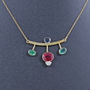 May include: A gold necklace with a curved bar and five dangling gemstones. The gemstones are red, green, and blue.
