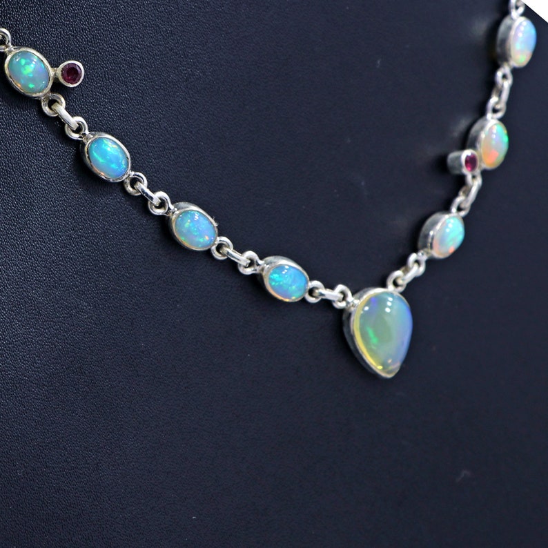 May include: A silver necklace with a teardrop-shaped opal pendant and smaller oval opals along the chain. The necklace also features small red gemstones.