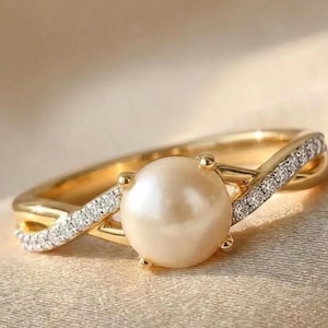 Op de afbeelding: Een gouden ring met een centrale ronde, crèmekleurige parel die door vier pootjes wordt vastgehouden. De band heeft een kruisend ontwerp met kleine heldere steentjes ingelegd langs de twee gouden armen die de parel omlijsten.