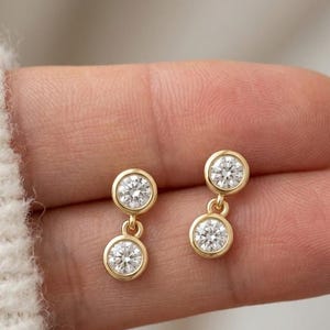 May include: A pair of gold drop earrings, each featuring two round, clear, faceted stones set in polished gold bezels. The top stone is fixed, while the bottom stone hangs from a small gold jump ring, creating a delicate, articulated design.
