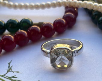 May include: A silver ring with a large, square-cut, light green gemstone. The ring is set against a backdrop of colorful beaded necklaces in shades of red, green, and cream, with a sprig of greenery in the foreground.