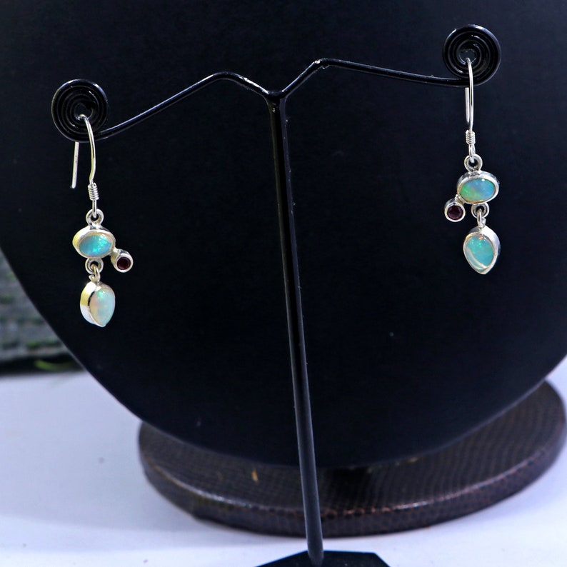 May include: A pair of silver dangle earrings with opal and garnet gemstones. The earrings feature a teardrop-shaped opal with a smaller opal and a garnet stone above it.