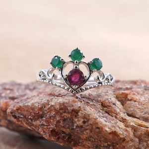 Puede incluir: Un anillo de plata con un diseño en forma de corazón que presenta una piedra roja rodeada de cuatro piedras verdes.