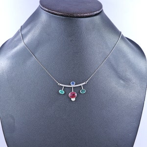 May include: A silver necklace with a bar pendant featuring a large oval red gemstone, a small blue gemstone, and two smaller green gemstones. The necklace is displayed on a black mannequin.
