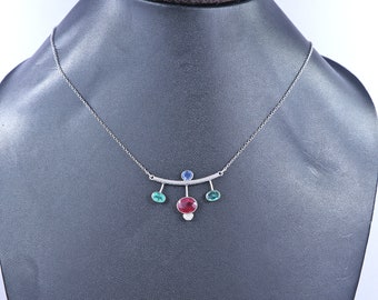 May include: A silver necklace with a bar pendant featuring a large oval red gemstone, a small blue gemstone, and two smaller green gemstones. The necklace is displayed on a black mannequin.