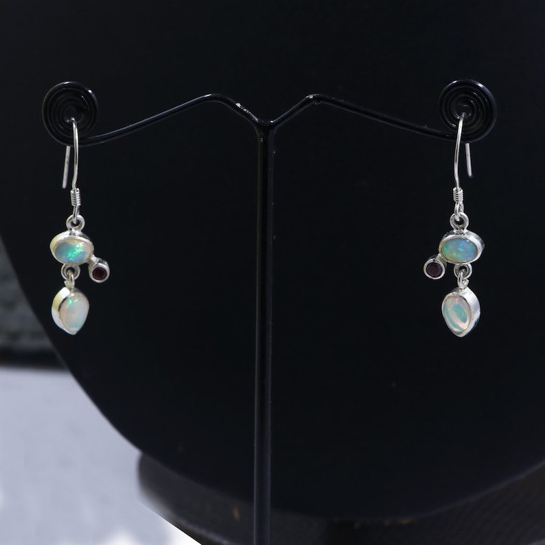 May include: A pair of silver dangle earrings with opal and garnet gemstones. The earrings are hanging from a black stand.