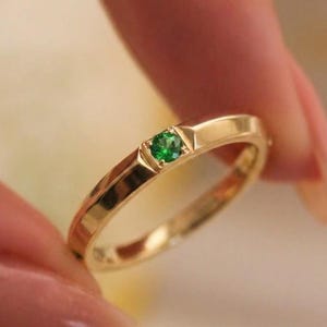 Puede incluir: Un anillo de banda dorada que presenta una pequeña piedra preciosa verde de corte redondo engastada al ras en el metal. El anillo tiene un acabado liso y pulido, con un diseño sencillo y limpio y un perfil plano en la parte superior central.