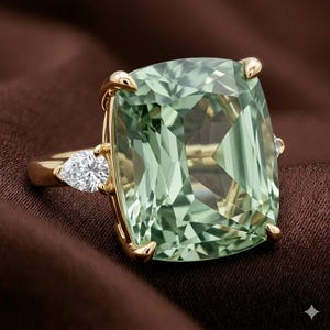 Op de afbeelding: Een gouden ring met een grote, rechthoekige, lichtgroene edelsteen. Aan de zijkant is een peervormige, heldere edelsteen gezet. De ring is geplaatst tegen een bruine achtergrond.
