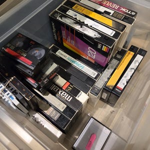 May include: A clear plastic storage bin filled with various VHS tapes and smaller Hi8 tapes. The tapes are in their original packaging, with labels featuring brands like TDK, Maxell, and Polaroid. Some labels are yellow, black, and white.