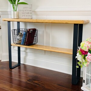 May include: A wooden console table with black metal legs and a shelf. The table has a natural wood finish and is decorated with white tulips in a glass vase, a wire basket with books, and a sign that reads "grow old along with me the best is yet to be..."