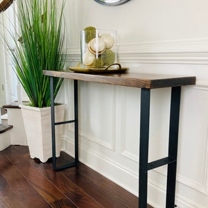 May include: A wooden console table with black metal legs. The table has a dark wood finish and is decorated with a vase of green plants and a glass jar filled with decorative balls.