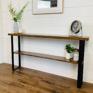 Puede incluir: Una mesa consola de madera marr&oacute;n oscuro con patas de metal negro y un estante. La mesa tiene un jarr&oacute;n blanco con flores y un reloj en la parte superior.