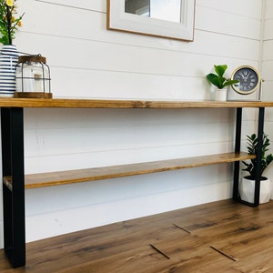 May include: A wooden console table with black metal legs and a shelf. The table has a natural wood finish and is 48 inches long. It is sitting on a wood floor.