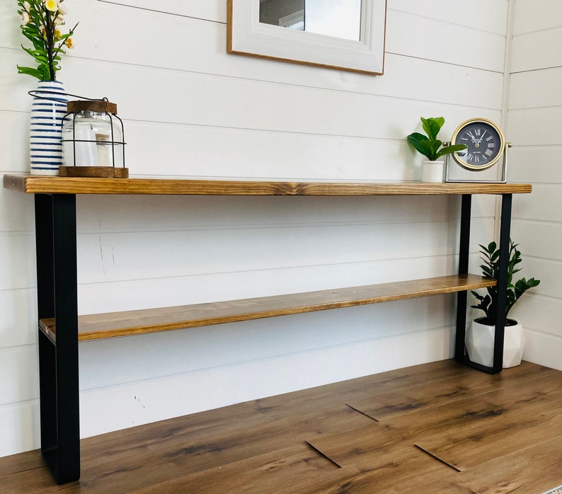 May include: A wooden console table with black metal legs and a shelf. The table has a natural wood finish and is 48 inches long. It is sitting on a wood floor.