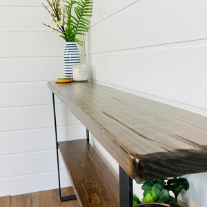 Puede incluir: Una mesa consola de madera con patas de metal negro y dos estantes. La mesa tiene un acabado de madera natural y est&aacute; decorada con un jarr&oacute;n de flores y una vela.
