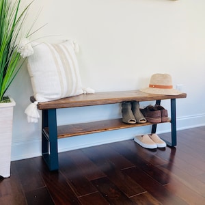 May include: A wooden bench with a black metal frame and a shelf underneath. The bench has a white and brown striped pillow on top and a brown hat and shoes on the shelf.
