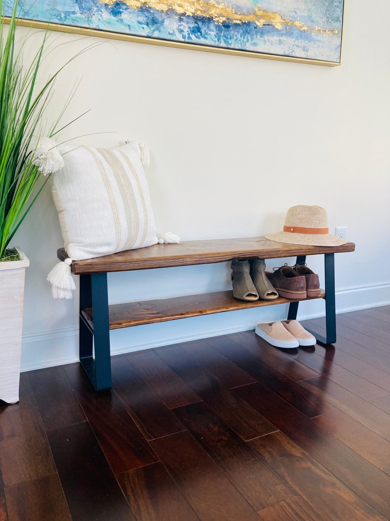 May include: A wooden bench with a black metal frame and a shelf underneath. The bench has a white and brown striped pillow on top and a brown hat and shoes on the shelf.