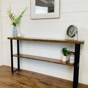 May include: A wooden console table with a black metal frame and a lower shelf. The table has a distressed wood finish and is decorated with a vase of flowers, a candle, a clock, and a potted plant.