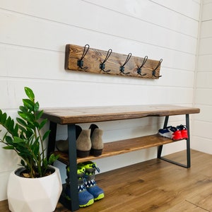 May include: A wooden bench with a metal frame and two shelves for storing shoes. The bench has a rustic brown finish and black metal legs. There are hooks on the wall above the bench.