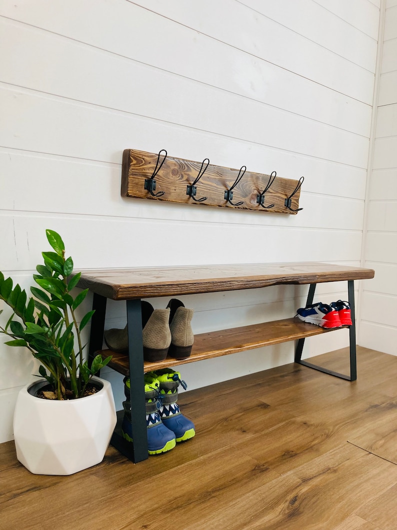 May include: A wooden bench with a metal frame and two shelves for storing shoes. The bench has a rustic brown finish and black metal legs. There are hooks on the wall above the bench.