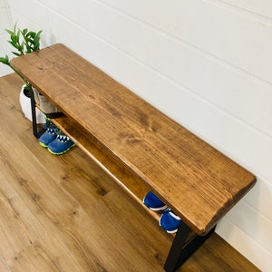 May include: A wooden bench with a black metal frame and a shelf underneath. The bench has a dark brown stain and is sitting on a light brown wood floor. There are two pairs of shoes on the shelf.