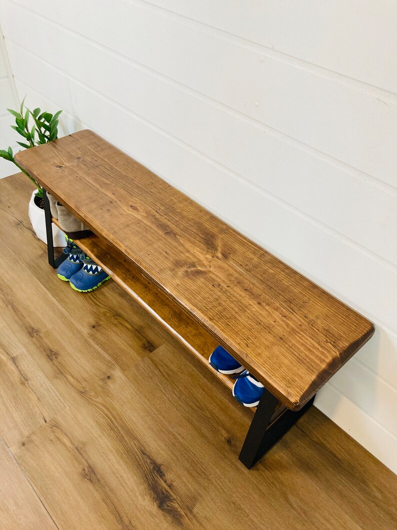 May include: A wooden bench with a black metal frame and a shelf underneath. The bench has a dark brown stain and is sitting on a light brown wood floor. There are two pairs of shoes on the shelf.