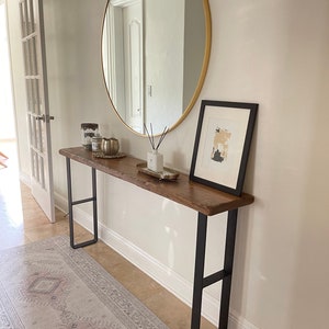 May include: A wooden console table with black metal legs. The table is decorated with candles, a gold bowl, and a diffuser. A round gold mirror hangs on the wall above the table. A framed abstract art print hangs on the wall to the right of the table.