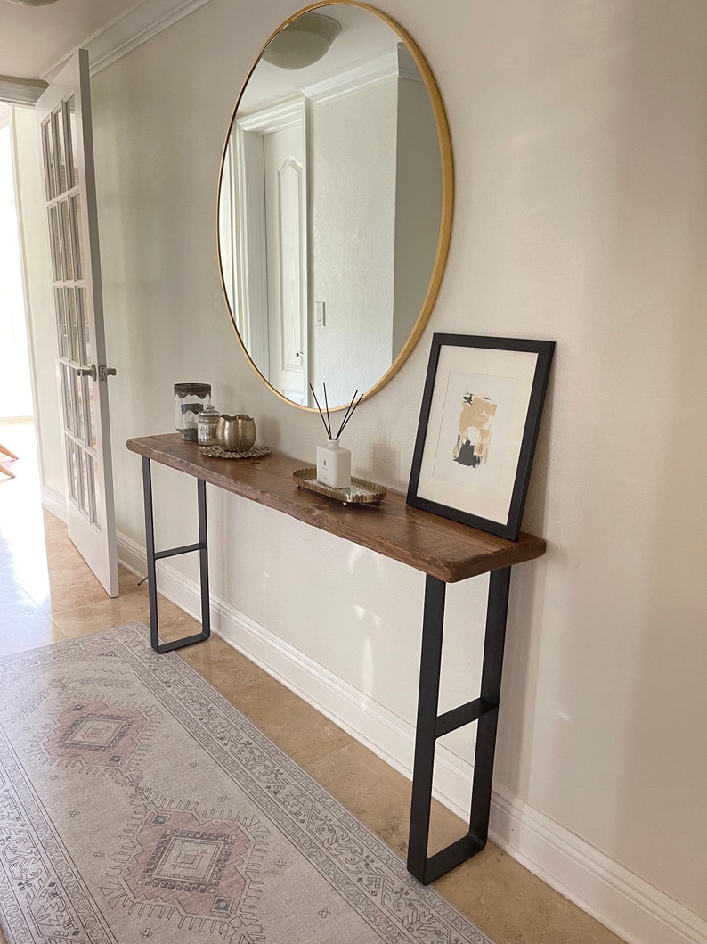 May include: A wooden console table with black metal legs. The table is decorated with candles, a gold bowl, and a diffuser. A round gold mirror hangs on the wall above the table. A framed abstract art print hangs on the wall to the right of the table.