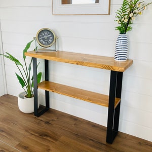 May include: A wooden console table with a black metal frame and two shelves. The table has a natural wood finish and is decorated with a clock and a vase of flowers.