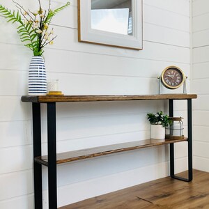 Puede incluir: Una mesa consola de madera con patas de metal negro y dos estantes. La mesa tiene un acabado de madera envejecida y est&aacute; decorada con un jarr&oacute;n de flores, un reloj y una planta en maceta.