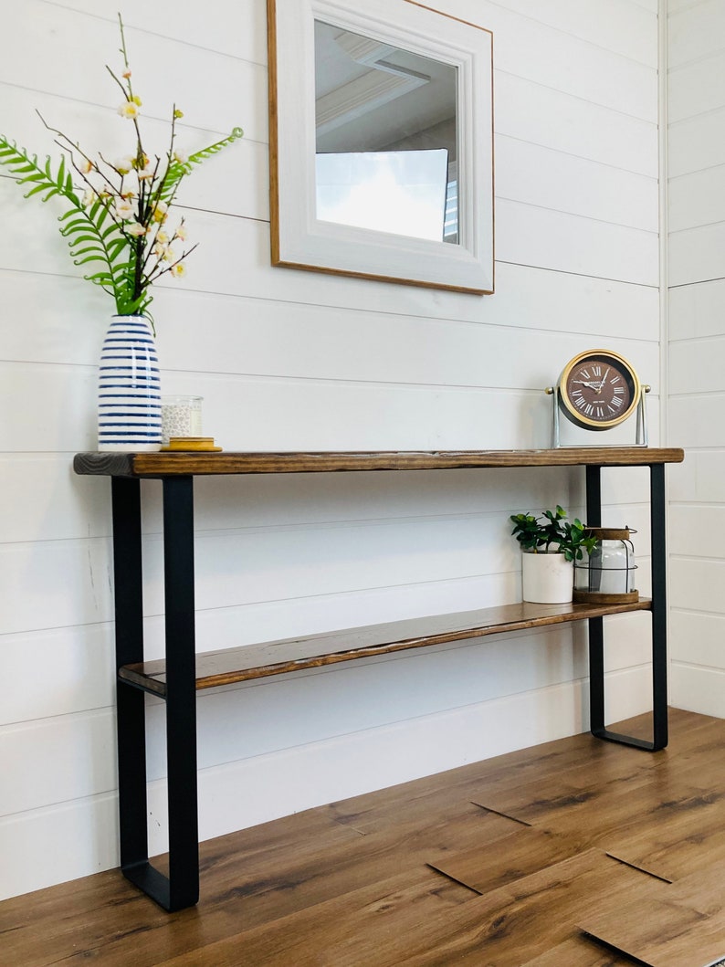 Puede incluir: Una mesa consola de madera con patas de metal negro y dos estantes. La mesa tiene un acabado de madera envejecida y est&aacute; decorada con un jarr&oacute;n de flores, un reloj y una planta en maceta.