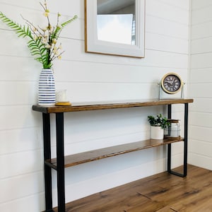 May include: A wooden console table with black metal legs and a shelf. The table has a distressed wood finish and is decorated with a vase of flowers, a candle, and a clock.