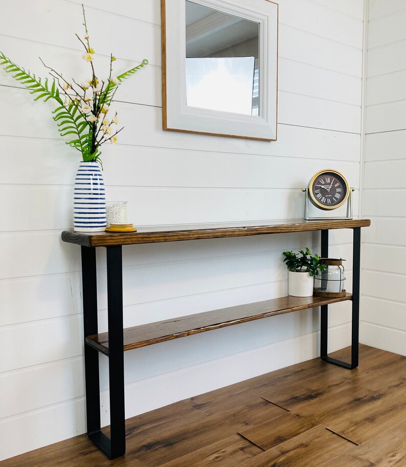 May include: A wooden console table with black metal legs and a shelf. The table has a distressed wood finish and is decorated with a vase of flowers, a candle, and a clock.