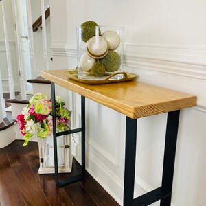 May include: A wooden console table with black metal legs. The table has a light wood finish and is decorated with a glass vase filled with green and white decorative balls.