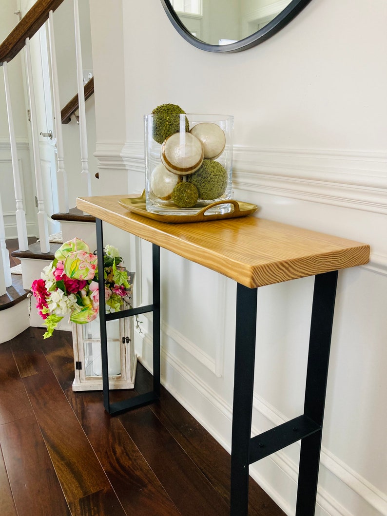 May include: A wooden console table with black metal legs. The table has a light wood finish and is decorated with a glass vase filled with green and white decorative balls.